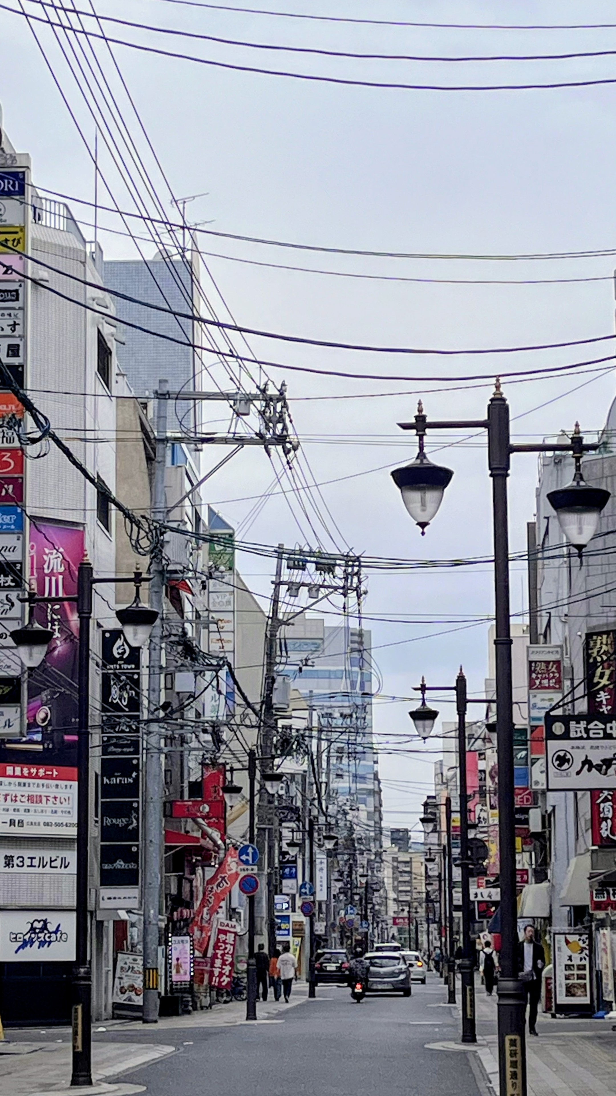 Eine Straßenszene mit Autos in der Ferne und einigen Menschen im Hintergrund, links und rechts mit sehr unterschiedlich hohen Fassaden. Sehr viel Kabel verlaufen in der Luft parallel und quer zur Straße. Im Vordergrund rechts ist ein Laternenpfahl mit je einer Laterne links und rechts sehr weit oben zu sehen.